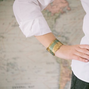 May include: A gold-toned bracelet with a map design. The bracelet is worn on a person's wrist.