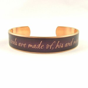 May include: A copper bracelet with a dark purple enamel band. The bracelet has the inscription "Our souls are made of, his and mine" in a copper-colored font.
