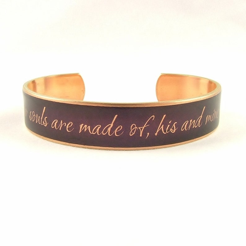 May include: A copper bracelet with a dark purple enamel band. The bracelet has the inscription "Our souls are made of, his and mine" in a copper-colored font.