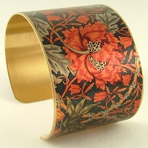 May include: A gold metal cuff bracelet with a floral pattern. The pattern features red, orange, and green flowers on a black background.