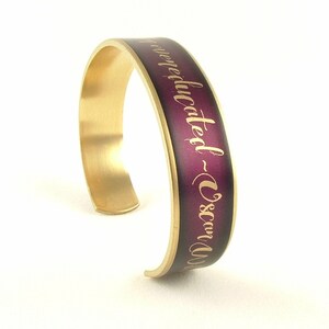 May include: Gold cuff bracelet with a purple gradient and the inscription "Never educated - Oscar Wilde" in gold lettering.