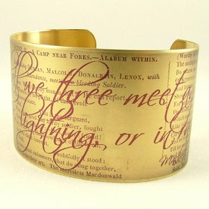 Peut inclure: Bracelet manchette doré avec un aspect vintage et vieilli. Le bracelet présente du texte bordeaux d'une pièce de théâtre, avec des phrases comme "free meet" et "lightning or in". Le bracelet est large et ouvert.