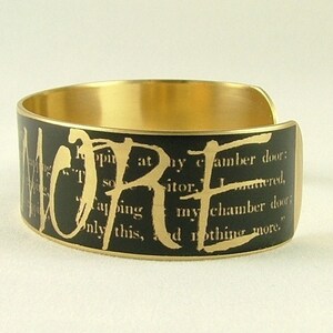 May include: A gold metal cuff bracelet with a black background and the word "LORE" printed in gold letters. The background is a black and white printed page with text.