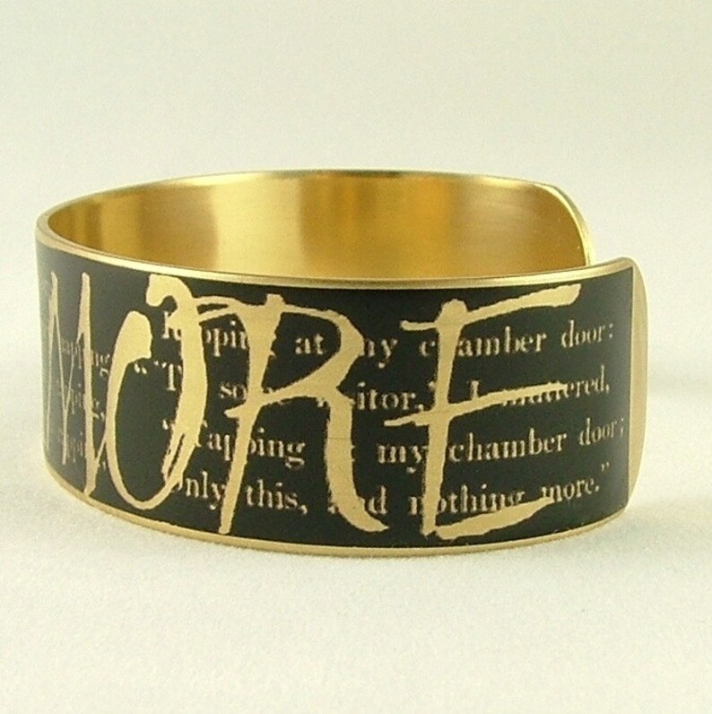 May include: A gold metal cuff bracelet with a black background and the word "LORE" printed in gold letters. The background is a black and white printed page with text.