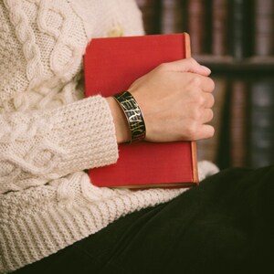 May include: A person wearing a white cable knit sweater holds a red book with a black and gold bracelet on their wrist. The bracelet has the words "VERA WANG" engraved on it.
