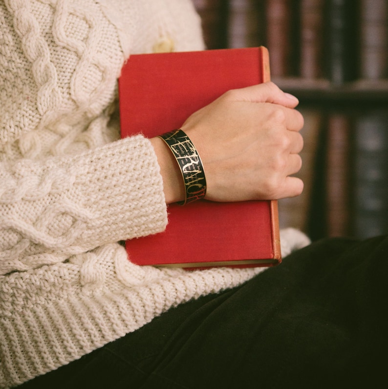 May include: A person wearing a white cable knit sweater holds a red book with a black and gold bracelet on their wrist. The bracelet has the words "VERA WANG" engraved on it.