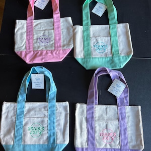 May include: Four mini pastel canvas tote bags. Each bag has a different color scheme: pink, mint green, light blue, and lavender. The bags have a cream-colored body with matching colored handles, bottom, and pocket. The Trader Joe's logo is embroidered on the front.