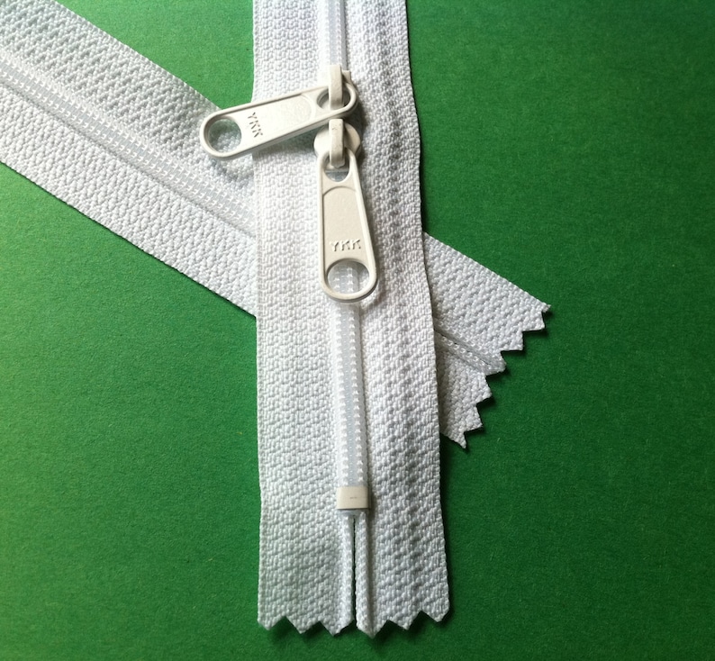 May include: White zipper with two white pull tabs. The zipper is closed and the pull tabs are facing each other. The zipper is on a green background.