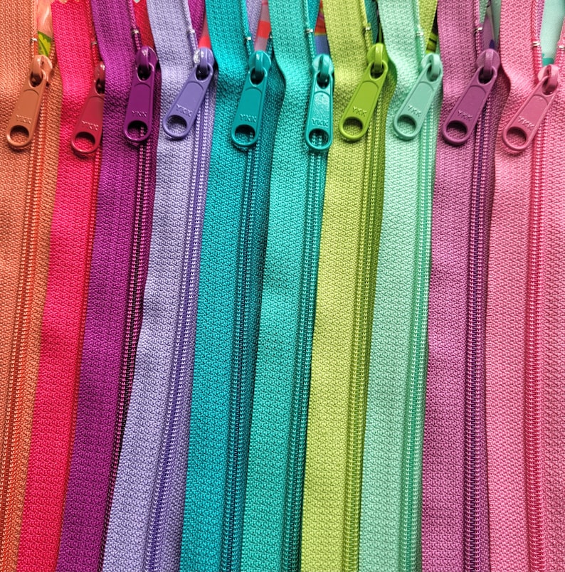 May include: A close-up of a variety of colourful zipper tapes. The zippers are arranged in a row, with the slider ends facing the viewer. The colours include red, purple, lavender, teal, green, and pink.