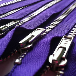 Op de afbeelding: Een reeks paarse ritsen met zilverkleurige metalen tanden en bijpassende zilverkleurige metalen schuivers. De stoffen band is diep, levendig paars van kleur en de metalen onderdelen hebben een gepolijste afwerking met zichtbare YKK-branding.