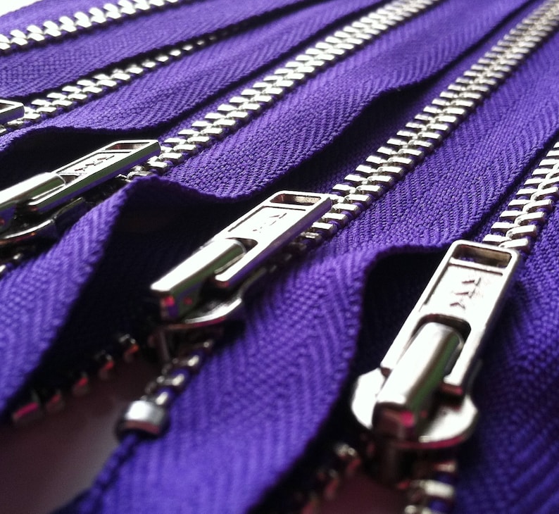 Op de afbeelding: Een reeks paarse ritsen met zilverkleurige metalen tanden en bijpassende zilverkleurige metalen schuivers. De stoffen band is diep, levendig paars van kleur en de metalen onderdelen hebben een gepolijste afwerking met zichtbare YKK-branding.