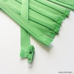 May include: A close-up of a bundle of green zippers with metal slider pulls. The zippers are laid out in a fan shape on a white surface.