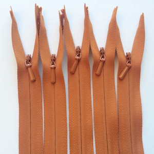 May include: Six brown zipper closures with metal sliders. The zippers are all closed and laid out in a row.