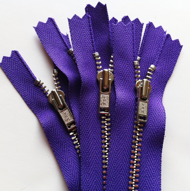 Op de afbeelding: Een verzameling van drie paarse ritsen met zilverkleurige metalen tanden en schuivers. Elke schuiver is duidelijk voorzien van het YKK-merklogo. De stoffen band is felpaars van kleur en de metalen onderdelen hebben een gepolijste afwerking.