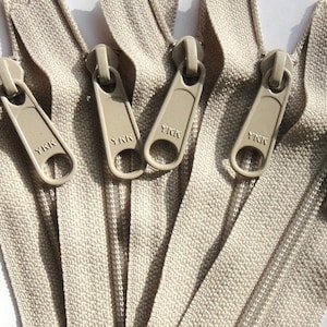 Può includere: Primo piano di una fettuccia di cerniera beige con cinque cursori YKK. I cursori sono in plastica e di colore beige.