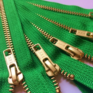 May include: A close-up of several green zipper tapes with gold metal teeth and sliders. The sliders have the word "YKK" printed on them.