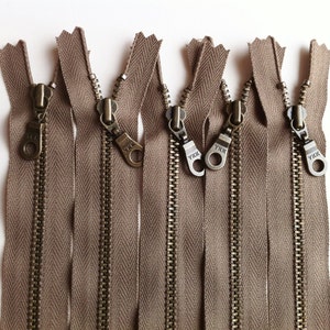 May include: Close-up of six brown zipper pulls with metal teeth and YKK branding. The zipper pulls are arranged in a row and are ready to be sewn into a garment.