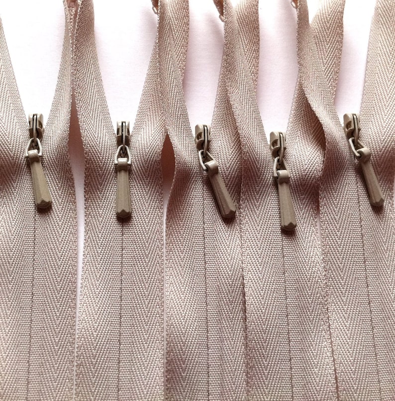 Op de afbeelding: Een close-up van zes beige ritsbanden met zilveren ritsen. De banden hebben een visgraatpatroon.