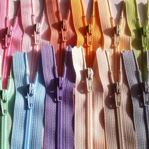 May include: Assortment of colorful zipper closures in various pastel shades, including green, blue, purple, pink, and orange. The zippers are arranged in a row, showcasing their different colors and styles.