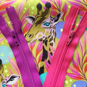 May include: Two zippers, one pink and one purple, are laid out on a colourful fabric with a giraffe print. The zippers are both closed and have metal slider pulls.