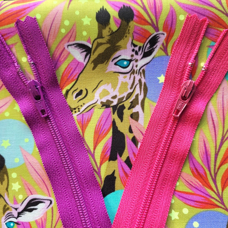 May include: Two zippers, one pink and one purple, are laid out on a colourful fabric with a giraffe print. The zippers are both closed and have metal slider pulls.