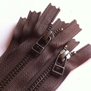 Puede incluir: Primer plano de una cremallera marrón con dos deslizadores metálicos. La cremallera está hecha de una tela texturizada y tiene un brillo sutil.