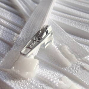 Puede incluir: Primer plano de un montón de cintas de cremallera blancas con un tirador de cremallera plateado. El tirador de la cremallera tiene una pequeña pieza de plástico en la parte inferior.
