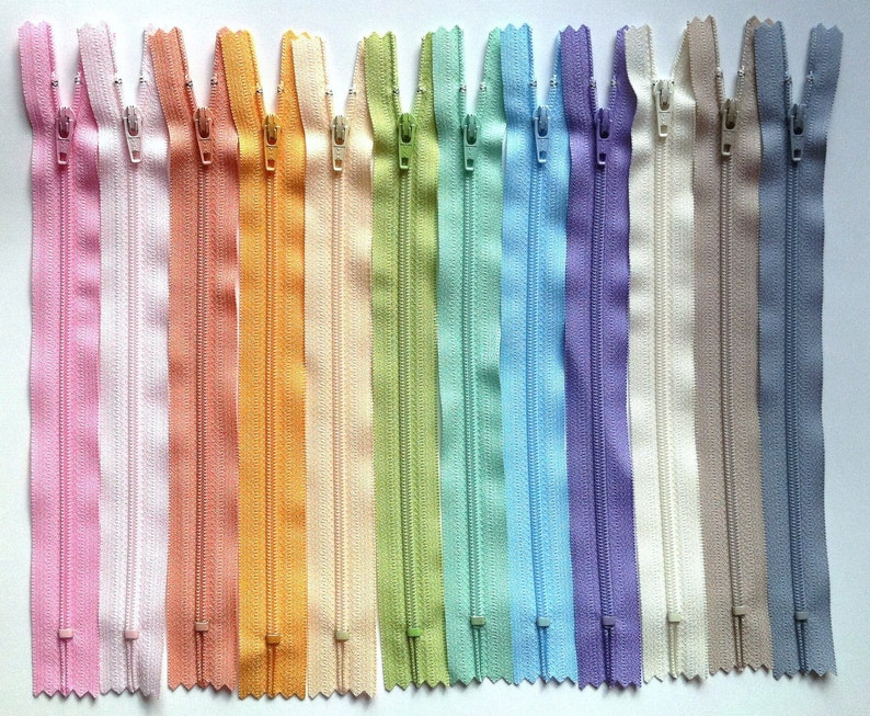 May include: A collection of 15 zipper tapes in various pastel colors, including pink, white, peach, orange, yellow, green, blue, purple, and beige. The zippers are all the same size and have a smooth, even texture.