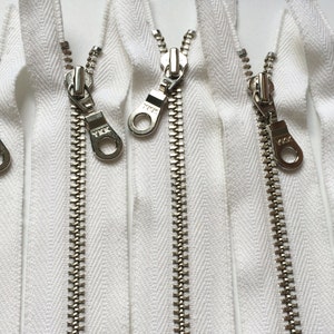 May include: Four white zipper tapes with silver metal teeth and pull tabs. Each pull tab has a silver loop and the text "YKK" printed on it.
