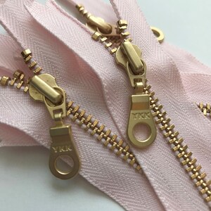 May include: Two gold zipper pulls with the YKK logo on a pink zipper tape.