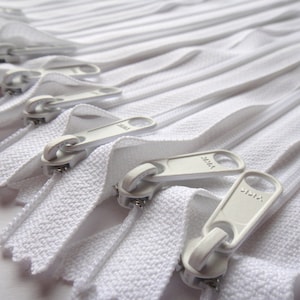 May include: A close-up of a pile of white zipper tapes with silver sliders. The zippers are all closed and the sliders are facing the camera.