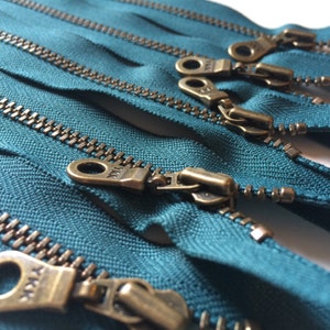 May include: Close-up of teal zipper tape with antique brass zipper pulls. The zipper tape is arranged in a stack, with the zipper pulls visible at the top of the stack.