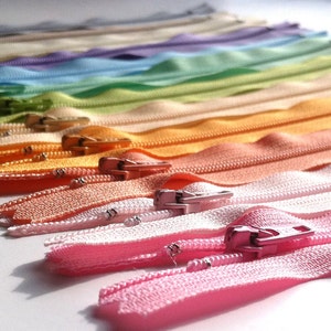 May include: A rainbow assortment of colorful zippers, arranged in a gradient from light beige to pink. The zippers are all closed and have a metal slider.