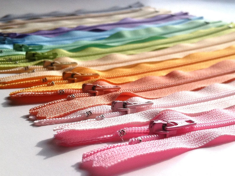 May include: A rainbow assortment of colorful zippers, arranged in a gradient from light beige to pink. The zippers are all closed and have a metal slider.