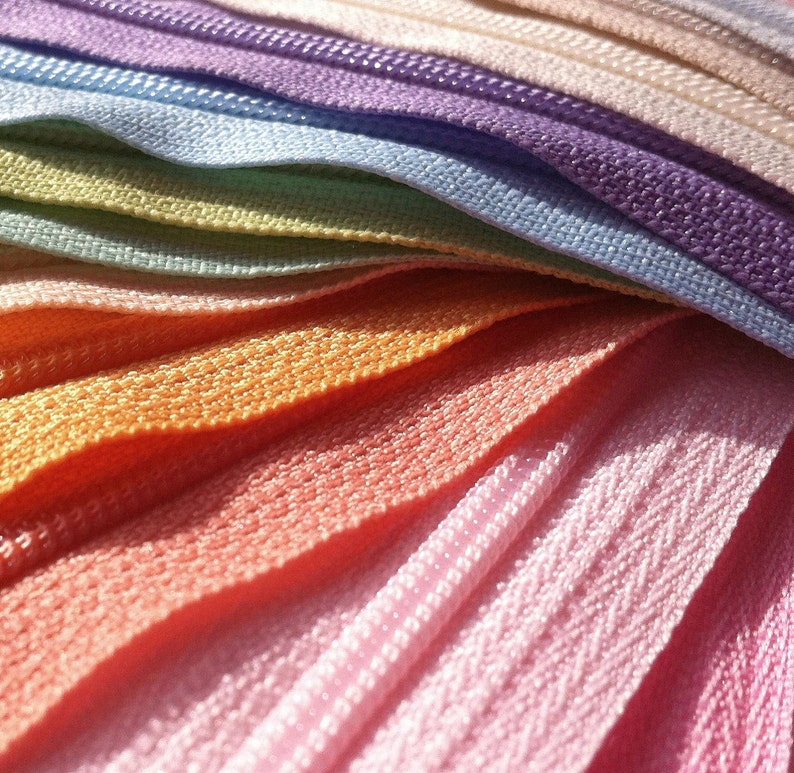 May include: A close-up of a rainbow assortment of zipper tape. The tape is arranged in a fan shape, with the colors transitioning from purple to pink to orange to yellow to green to blue.