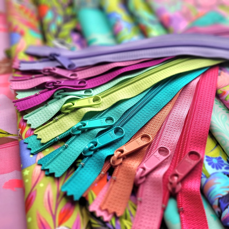 May include: A collection of colourful zippers in various shades of pink, green, blue, and orange. The zippers are arranged in a fan shape and are lying on top of a patterned fabric.