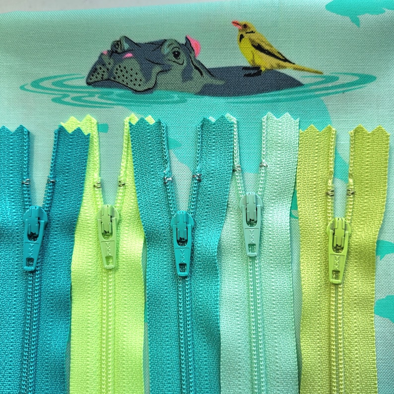 May include: A close-up of seven different zipper pulls in various shades of green and blue. The zipper pulls are arranged in a row, with the zipper teeth facing the viewer. The zipper pulls are made of metal and have a smooth, polished finish.