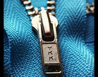 Op de afbeelding: Een close-up van een zilveren rits-trekker met de letters "YKK" erop gegraveerd. De rits zit op een felblauwe stof.