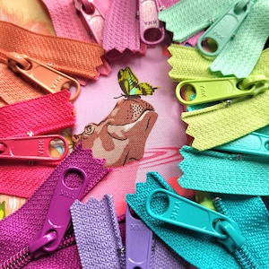 May include: A close-up of a variety of colourful zipper pulls with the brand name "YKK" printed on them. The zipper pulls are arranged in a circle around a fabric background with a hippopotamus and butterfly design.