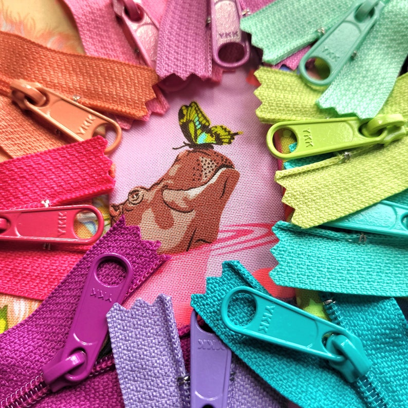 May include: A close-up of a variety of colourful zipper pulls with the brand name "YKK" printed on them. The zipper pulls are arranged in a circle around a fabric background with a hippopotamus and butterfly design.