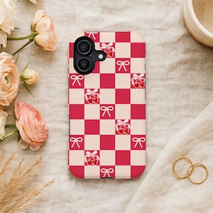 May include: A phone case with a red and cream checkerboard pattern. The case features a repeating design of red bows and cherries. The phone case is surrounded by flowers, a small ceramic cup, and two gold rings.