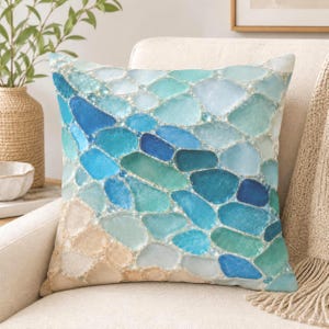 May include: A square throw pillow featuring a printed pattern of smooth, rounded sea glass pieces in shades of blue, teal, and white, transitioning into a sandy beige color at the bottom edge, creating a mosaic-like appearance.