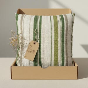 May include: A square decorative pillow with vertical stripes in shades of green, beige, and gray, inside a cardboard box. A small, dried floral arrangement and a blank tag are included. The pillow is a home decor item.