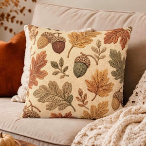 May include: A square throw pillow featuring a cream-colored background with a woven pattern of autumn leaves in shades of orange, brown, and green, interspersed with detailed illustrations of brown and green acorns.