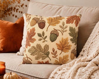 May include: A square throw pillow featuring a cream-colored background with a woven pattern of autumn leaves in shades of orange, brown, and green, interspersed with detailed illustrations of brown and green acorns.