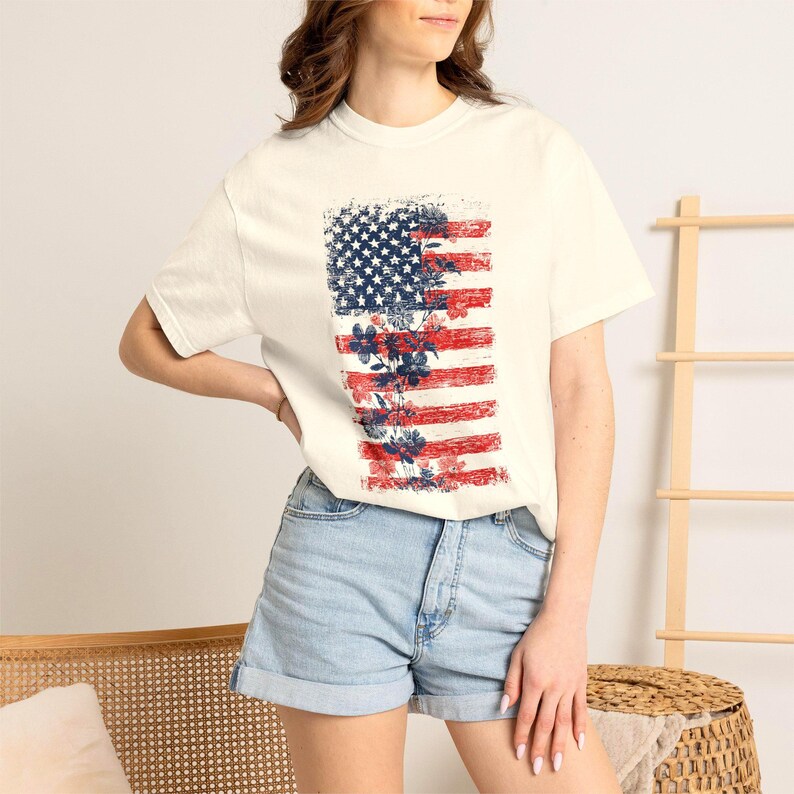 May include: Cream-coloured t-shirt with a distressed American flag design and floral accents. The flag's stars are blue, and the stripes are red. The shirt is worn with denim shorts, creating a casual, patriotic style.