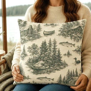 May include: A square throw pillow featuring a dark green toile-style print on a cream background. The pattern depicts a wilderness scene with log cabins, pine trees, a canoe on the water, swimming fish, and a duck floating on a lake.