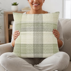 May include: A square throw pillow featuring a plaid pattern in shades of sage green, olive, and cream. The design consists of intersecting horizontal and vertical lines of varying thickness, creating a textured, woven appearance on the fabric surface.