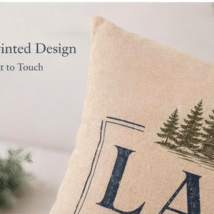 May include: A beige throw pillow cover featuring a printed design of a cabin and pine trees with the text Printed Design Flat to Touch visible on the left side. The fabric has a visible woven texture and the graphic is in dark blue and green.