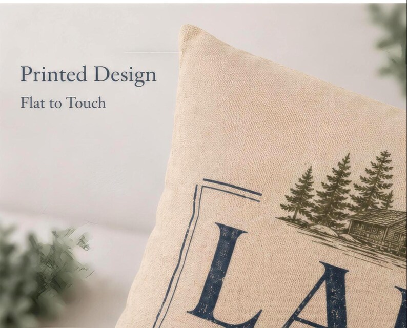 May include: A beige throw pillow cover featuring a printed design of a cabin and pine trees with the text Printed Design Flat to Touch visible on the left side. The fabric has a visible woven texture and the graphic is in dark blue and green.